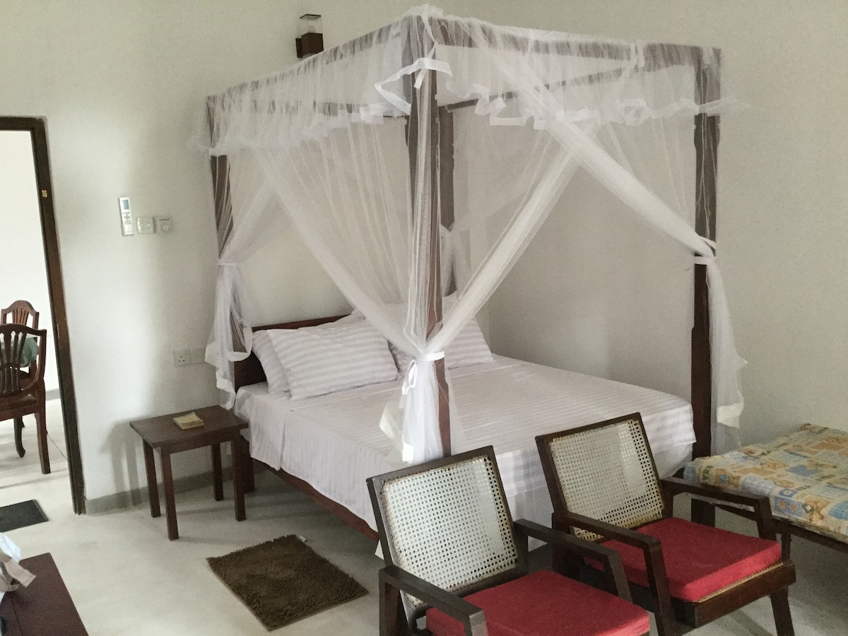 A spacious bedroom is presented with a four-poster bed draped in sheer white canopy fabric. Two armchairs with woven backs are positioned nearby. Soft lighting creates a calming environment, and the room features clean, minimalist decor.