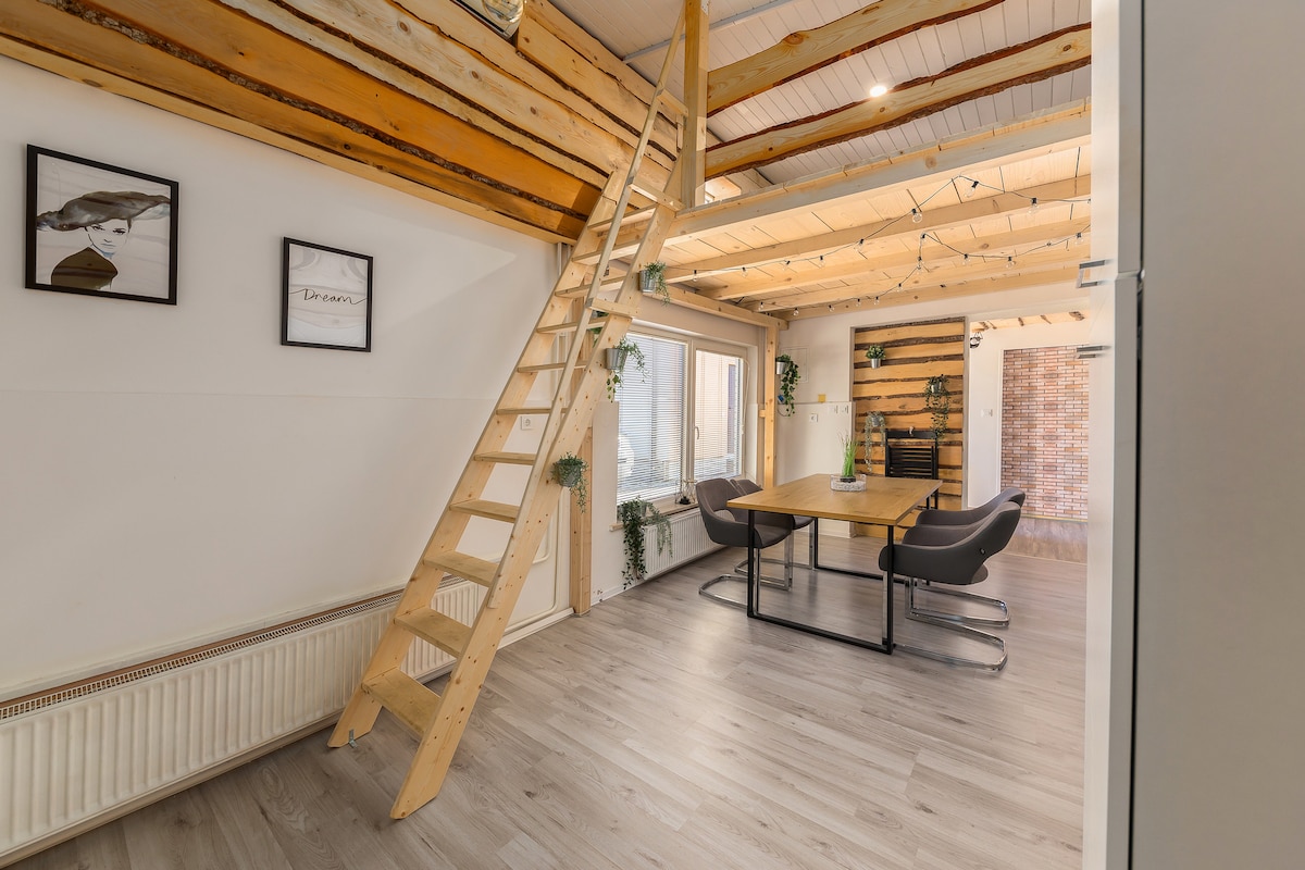 A spacious living area is highlighted by wooden beams on the ceiling and large windows allowing natural light to fill the space. A wooden table with chairs is visible, alongside a staircase leading to an upper loft area. Green plants add a touch of nature.