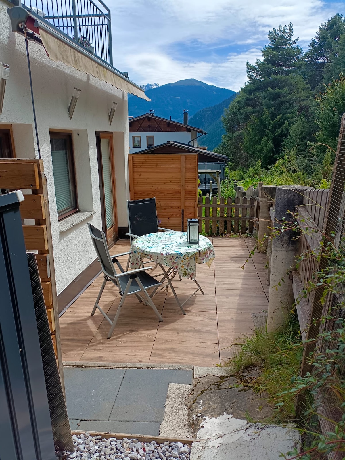 A private terrace is visible, featuring a small table and two chairs on a tiled surface. Surrounding greenery creates a natural setting, with a view of the mountains in the background. A wooden fence borders the space, enhancing privacy.