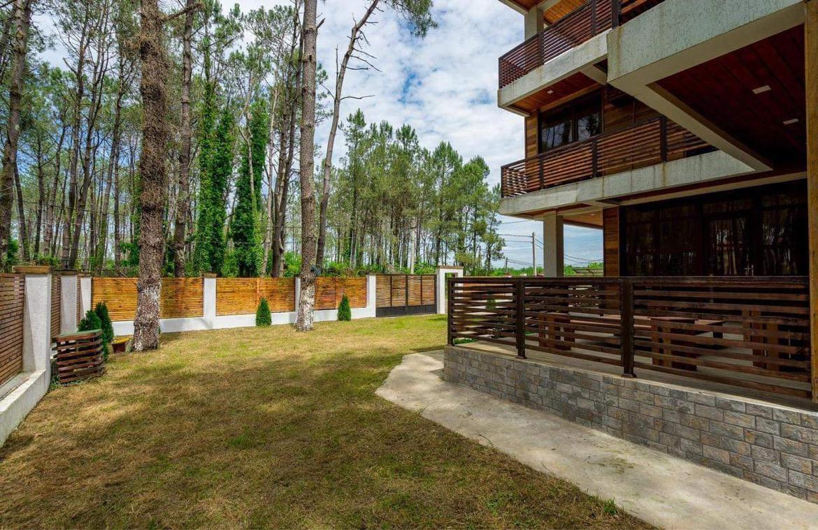 A spacious outdoor area surrounds the house, featuring a well-maintained lawn enclosed by wooden fencing. Several tall pine trees provide shade, and a small seating area is visible on the terrace. The building's wooden balconies overlook the serene landscape.