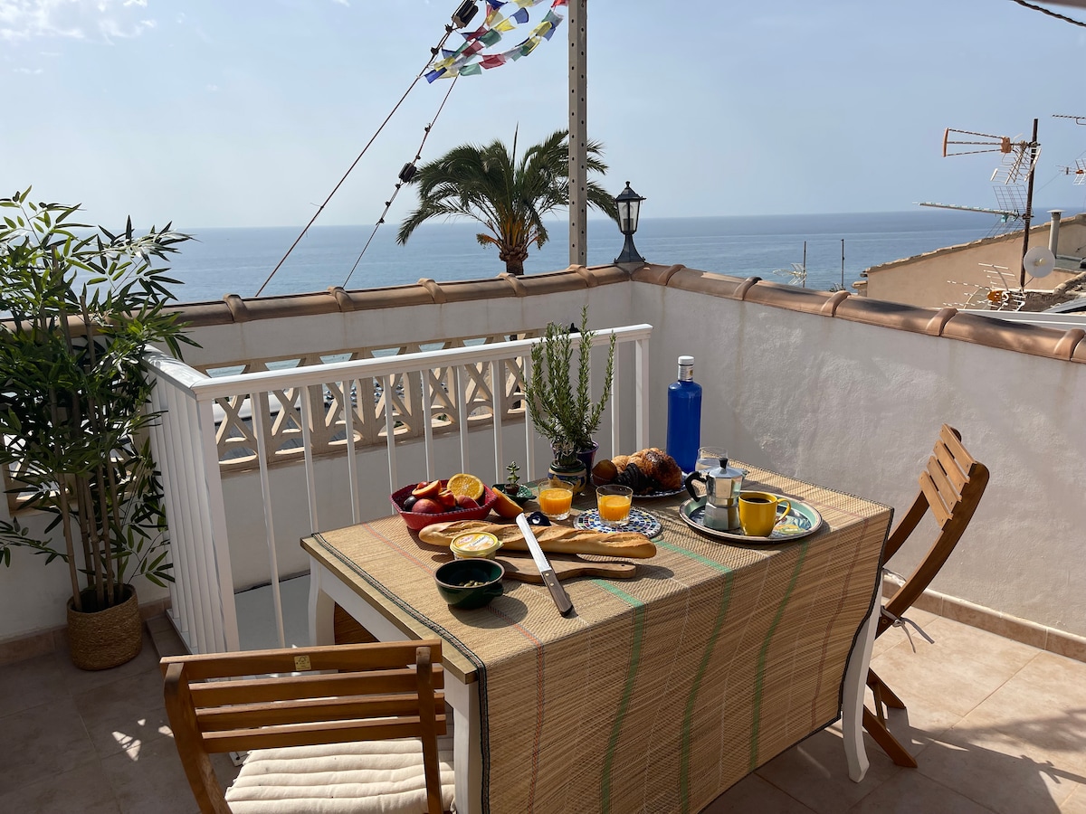 A terrace table is set for breakfast, featuring fresh fruit, drinks, and a serving tray. The view includes distant sea horizons and colorful fabric flags fluttering in the breeze. Surrounding greenery adds a touch of nature to the space.