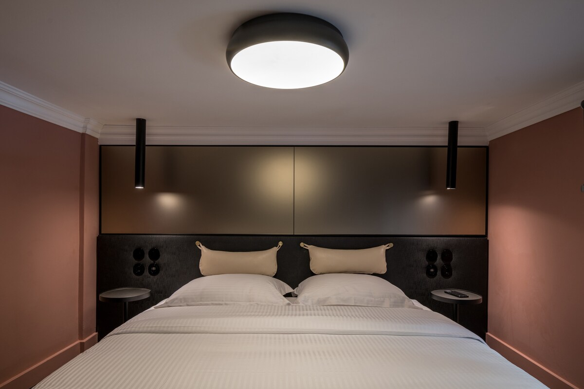 The refined sleeping area features a spacious double bed with upholstered headboard and adjacent modern bedside tables. Soft lighting from overhead fixtures creates a soothing ambiance, while warm walls complement the minimalist design.