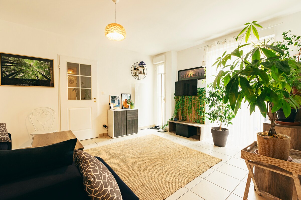 The living area features natural light streaming through large windows, illuminating the space. A cozy seating arrangement is complemented by a jute rug. Green plants are positioned around the room, adding a touch of nature, while a decorative wall piece provides visual interest.
