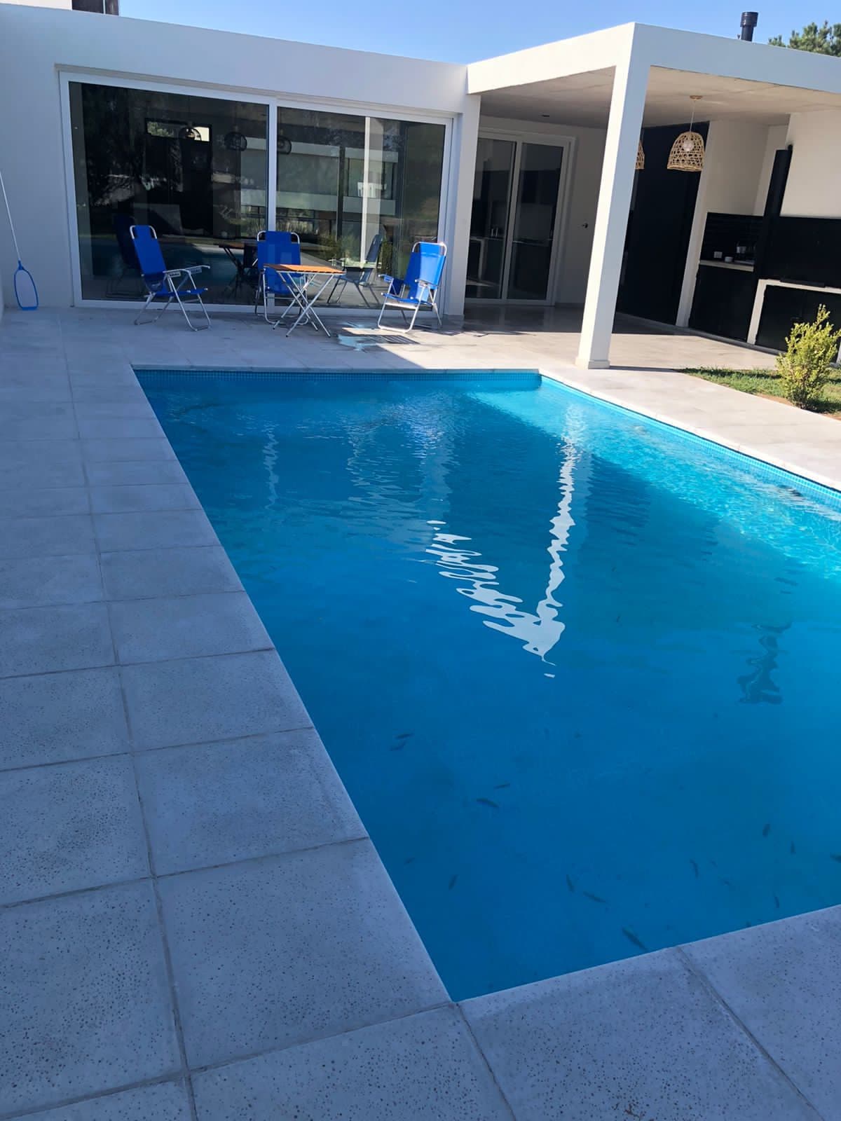 A clear blue pool reflects sunlight in a spacious outdoor area. Two blue chairs and a small table are positioned nearby, creating a relaxing spot for outdoor dining or lounging. Large glass doors provide a view into the interior space.