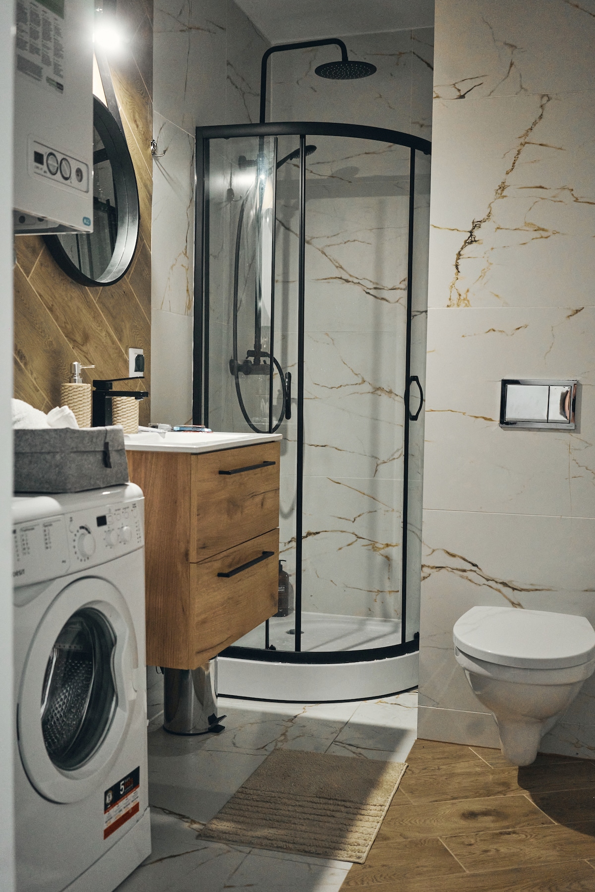 A compact bathroom features a glass shower stall with a modern showerhead. A washing machine is situated near a wooden cabinet with drawers. Light-colored tiles and a round mirror contribute to the overall bright and functional design.
