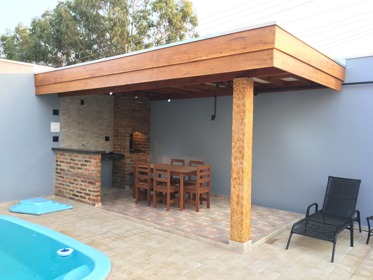 An outdoor area features a rustic wooden structure with a spacious dining table surrounded by six wooden chairs. A small stone bar area is visible, adjacent to a pool with a light blue edge, while a sleek black lounge chair sits nearby.
