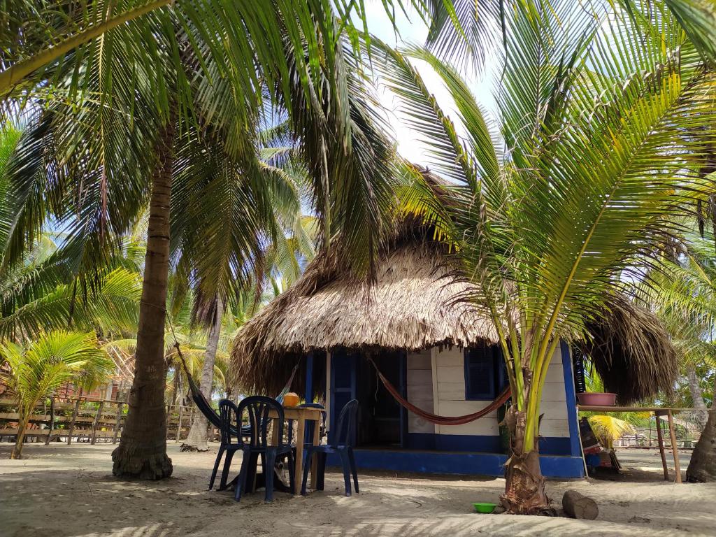 A charming cabin with a thatched roof is nestled among palm trees on a sandy beach. A small outdoor table with chairs is positioned nearby, offering a space for relaxation. The overall setting highlights a serene and tropical environment.