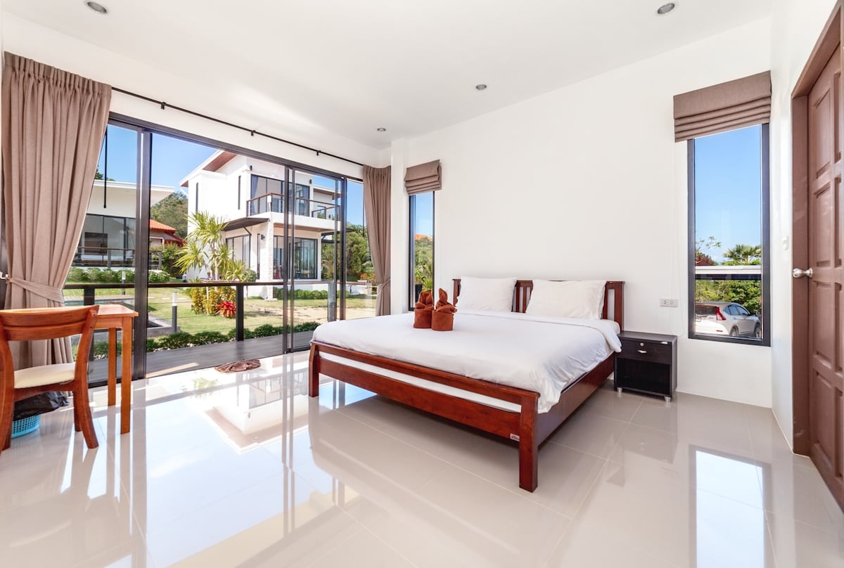 A light-filled room features a spacious bed with neutral-colored bedding, complemented by dark wood furnishings. Large glass windows provide views of the landscaped surroundings, and sheer curtains allow natural light to brighten the space. A wooden table and chair occupy one corner.
