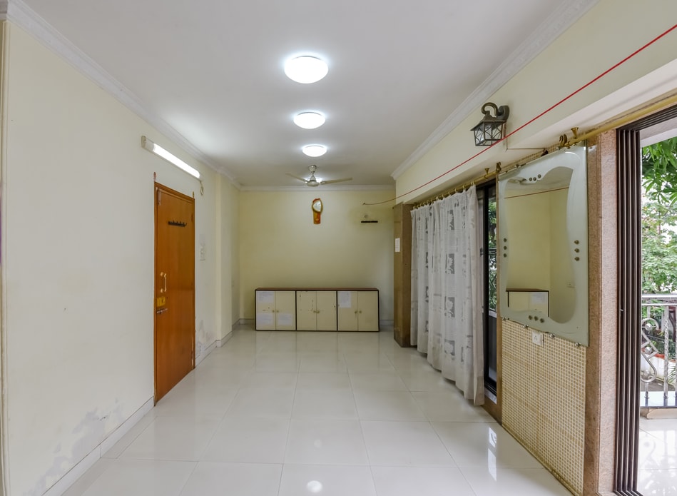 A spacious living area features light-colored walls and a tiled floor. Natural light is allowed through large windows that are adorned with sheer curtains. A mirror and a cabinet are positioned along one wall, while a ceiling fan hangs centrally.