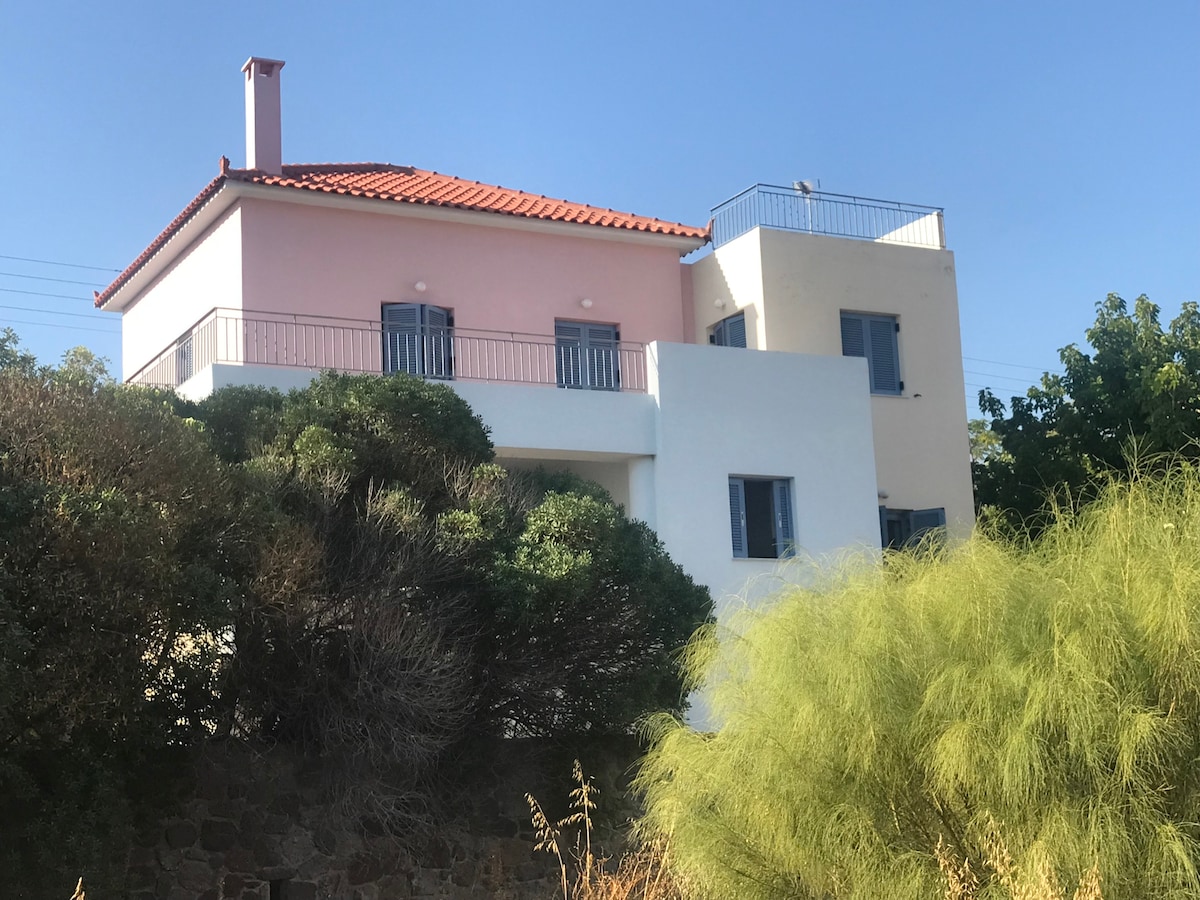 The two-story vacation house displays a combination of pink and white exterior walls, complemented by a tiled roof. Large windows provide natural light, while a balcony on the upper level offers outdoor seating. Surrounding greenery and shrubs enhance the property’s inviting appearance.