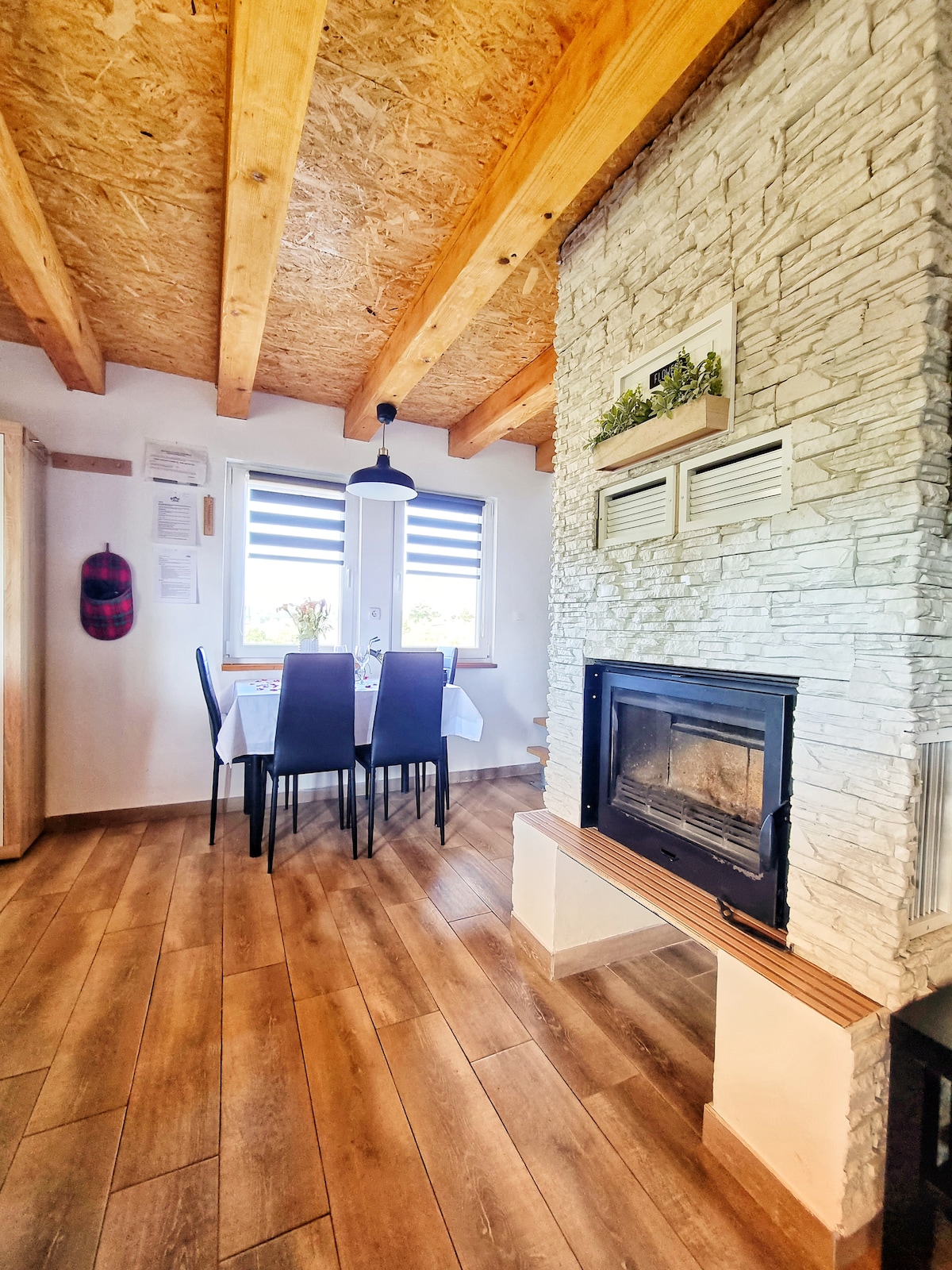 A dining area is illuminated by natural light from the windows, featuring a simple table with four dark chairs. A stone fireplace is integrated into the wall, complemented by wooden accents on the ceiling and floor, creating a cozy and functional space.