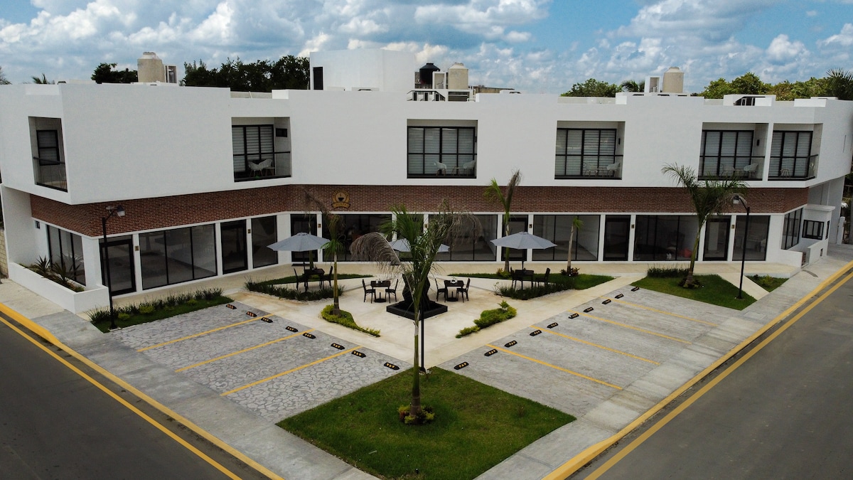 An aerial view of a modern building features a landscaped courtyard with palm trees and shaded seating areas. The facade showcases large windows and a mix of brick and white walls. A well-maintained parking area is visible, accommodating several vehicles.