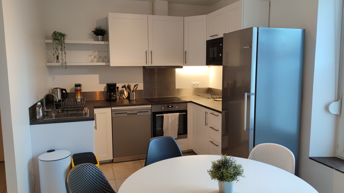 A modern kitchen area is displayed, featuring sleek cabinets and stainless steel appliances. A round dining table is surrounded by colorful chairs, with a small potted plant at its center. Essential kitchen amenities include a dishwasher, refrigerator, and countertop space for meal preparation.