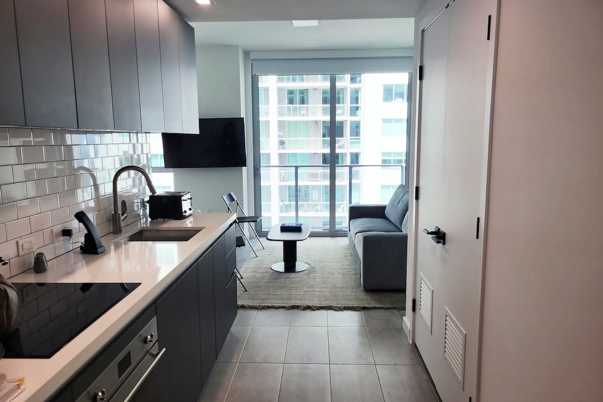A modern kitchen area featuring sleek gray cabinetry and stainless steel appliances is visible, complemented by a white countertop. A cozy seating area is shown with a gray sofa and small round table. Large windows allow natural light to fill the space, offering views of the city.