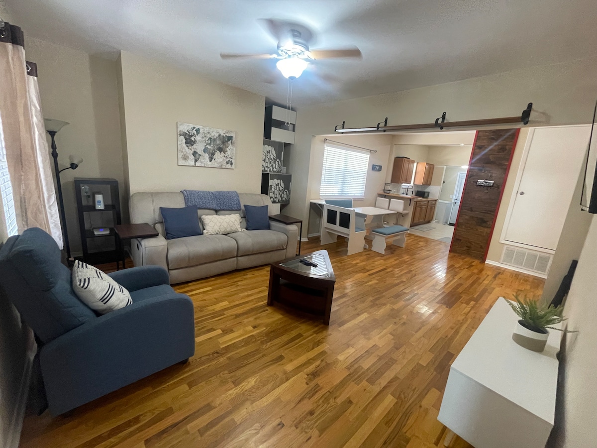 A living area features a light hardwood floor, with a comfortable sofa and armchair arranged around a coffee table. A ceiling fan provides airflow, while natural light enters through the windows. A TV stand and wall art add character to the space.