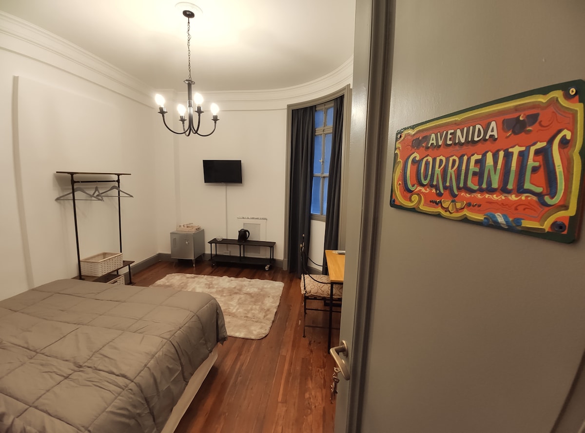 A spacious room features a warm wooden floor and a textured area rug. A comfortable bed is positioned against the wall. A wall-mounted television is visible across from a small table with chairs. A stylish chandelier hangs from the ceiling, enhancing the ambiance.