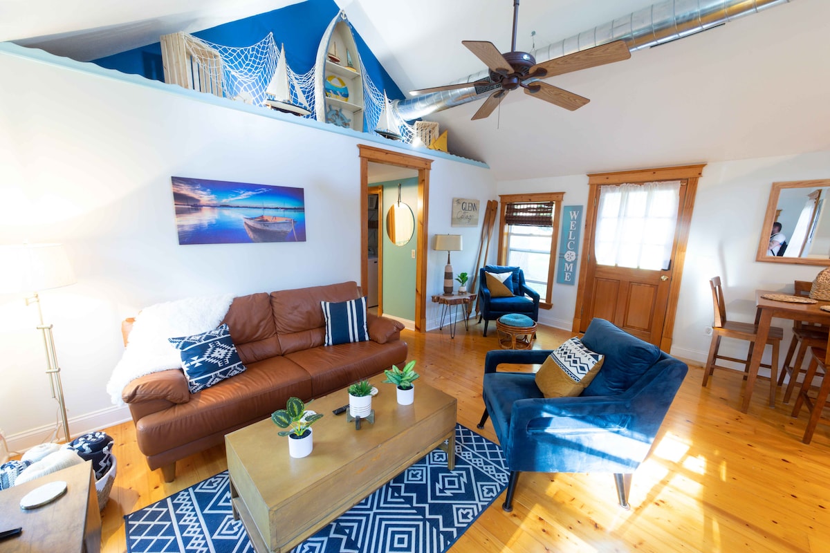 The living room features a brown leather couch and a blue accent chair, complemented by a wooden coffee table adorned with small plants. A geometric area rug lays underneath, and natural light streams in through a window, highlighting a welcoming atmosphere with rustic wood accents.