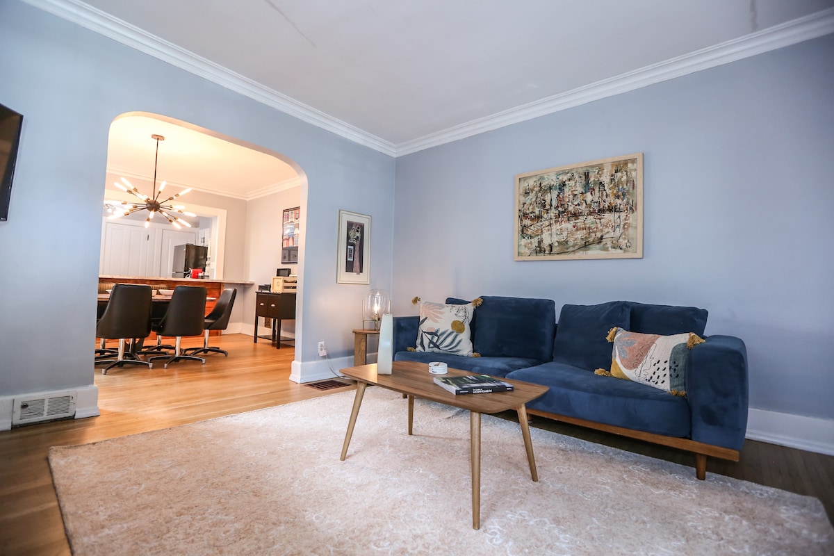 A cozy living room features a blue sofa adorned with decorative pillows. A wooden coffee table sits atop a light area rug. An archway leads to a dining area, with a view of modern light fixtures and dining chairs in the background.
