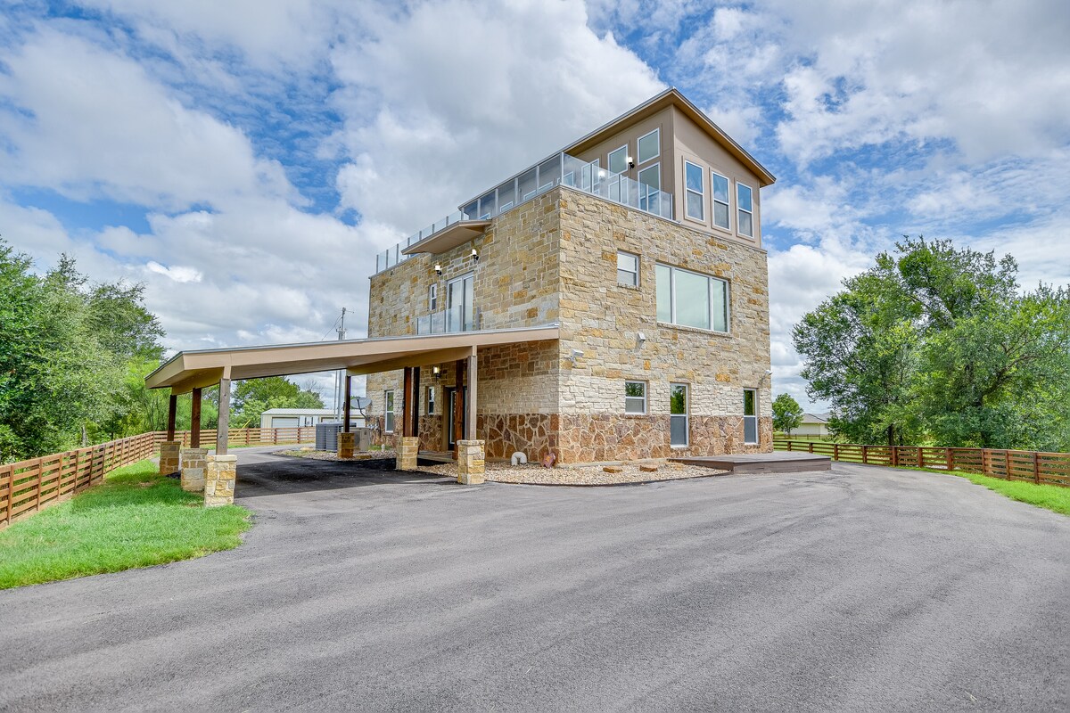 A modern three-story house with a blend of stone and stucco exterior is depicted. An expansive parking area leads to a covered entryway. A spacious deck is visible on the upper level, providing views of the surrounding landscape, with lush greenery and a clear sky in the background.