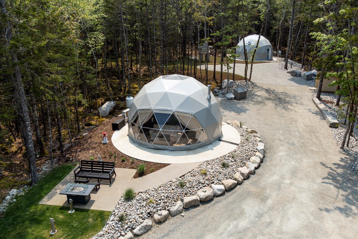 An aerial view of the geo-dome property reveals three domes set within a forested area. Surrounding greenery is complemented by a gravel pathway, a fire pit, and a bench. Natural stones line the perimeter, emphasizing the peaceful woodland setting.
