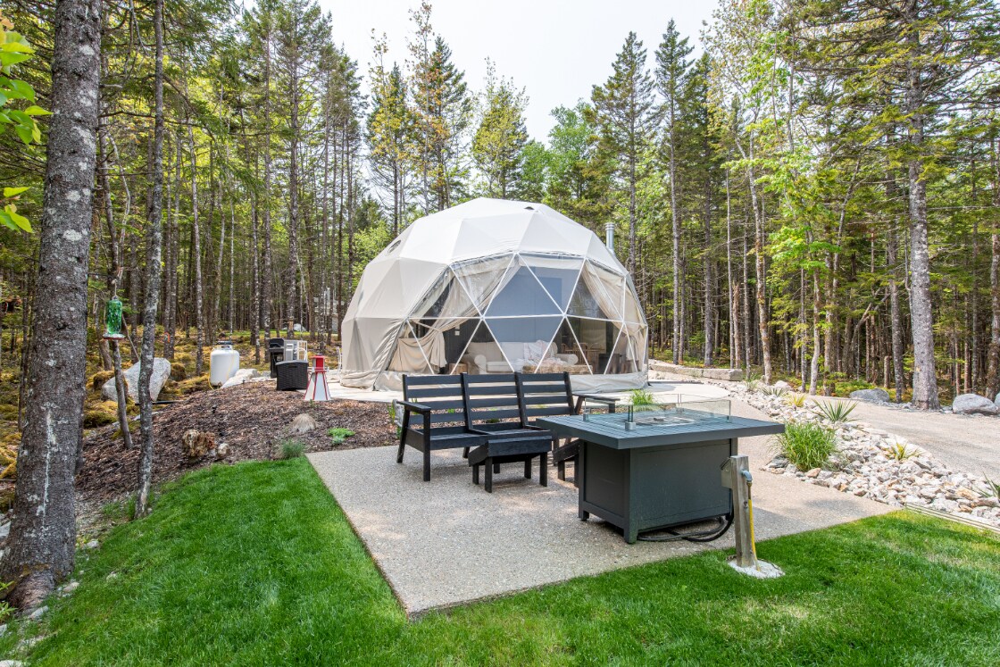 A spacious geo-dome is surrounded by lush trees, featuring large windows that provide a view of nature. An outdoor seating area is arranged on a patio, complemented by a fire pit and a barbecue, creating an inviting space for relaxation.