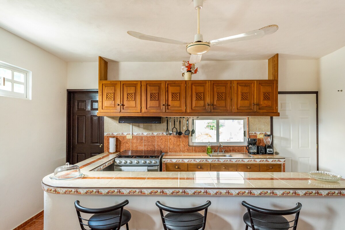 A functional kitchen features wooden cabinetry and a tiled countertop. An island with bar stools provides a casual dining area. Kitchen appliances, including a stove and coffee maker, are organized within easy reach, complemented by ample natural light from windows.