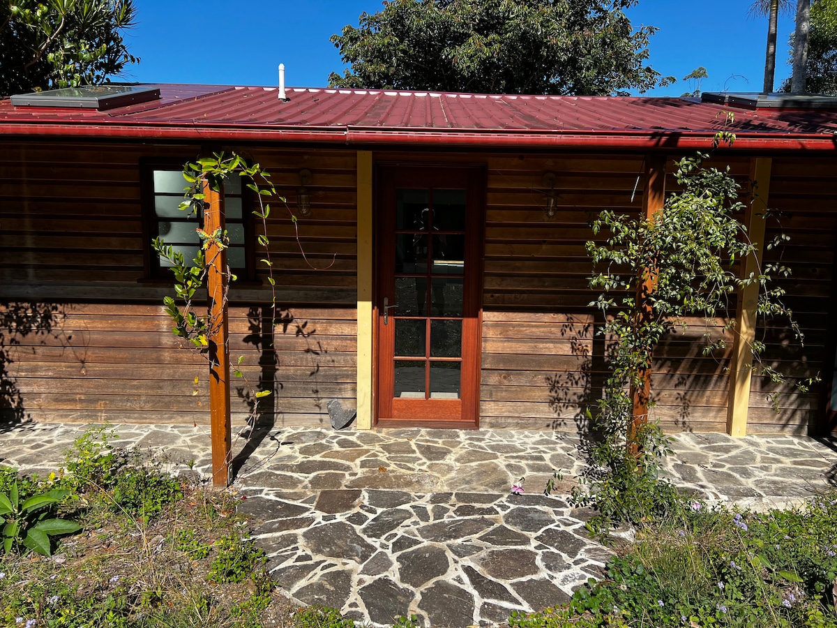 The exterior of a rustic cabin is highlighted, featuring wooden siding and a red metal roof. A stone pathway leads to the cabin's door, framed by greenery and flowering plants, creating a welcoming entryway.