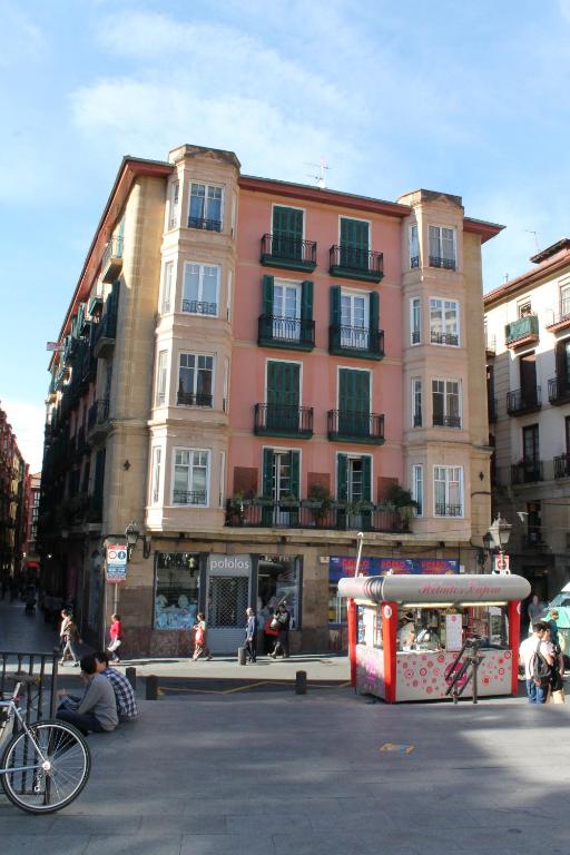 A multi-story building displays a combination of pink and beige façades, adorned with green shutters on the windows. Pedestrians are seen in the foreground, alongside a colorful kiosk and bicycles parked nearby, all set against a backdrop of a clear blue sky.