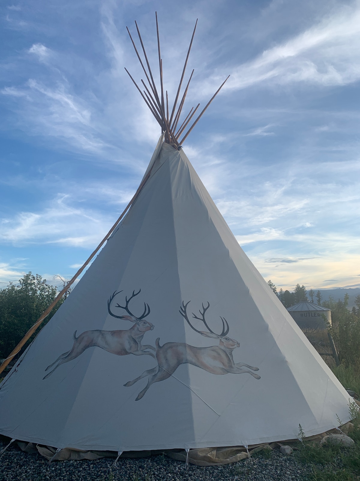A traditional tipi is showcased against a dusky sky, featuring artwork of two leaping deer on its exterior. The structure's conical shape is accentuated by tall wooden poles extending from the top. Surrounding greenery softly frames the scene, enhancing the natural setting.