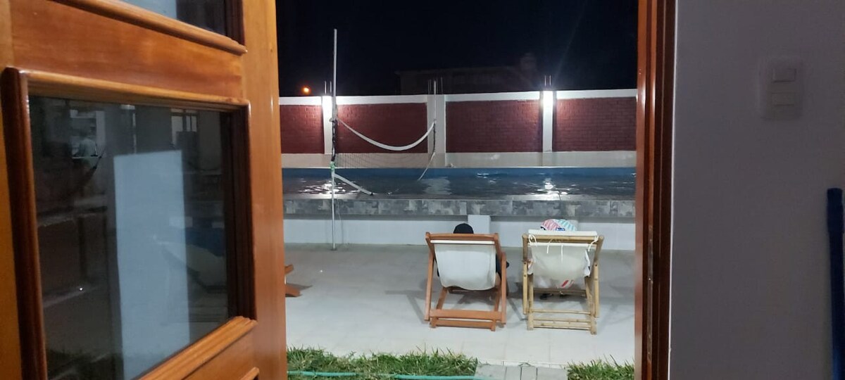 The image shows a view from an open door into an outdoor area featuring a spacious pool surrounded by a decorative wall. Two lounge chairs are positioned nearby, providing a relaxed space for seating under nighttime illumination.