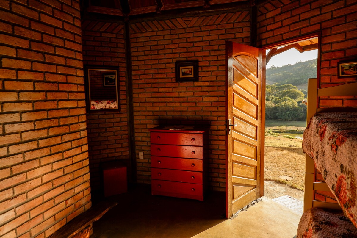A rustic room features a brown brick wall complemented by a wooden door, which opens to a scenic view. A red dresser with multiple drawers sits against the wall, and sunlight filters in, highlighting the warmth of the space.