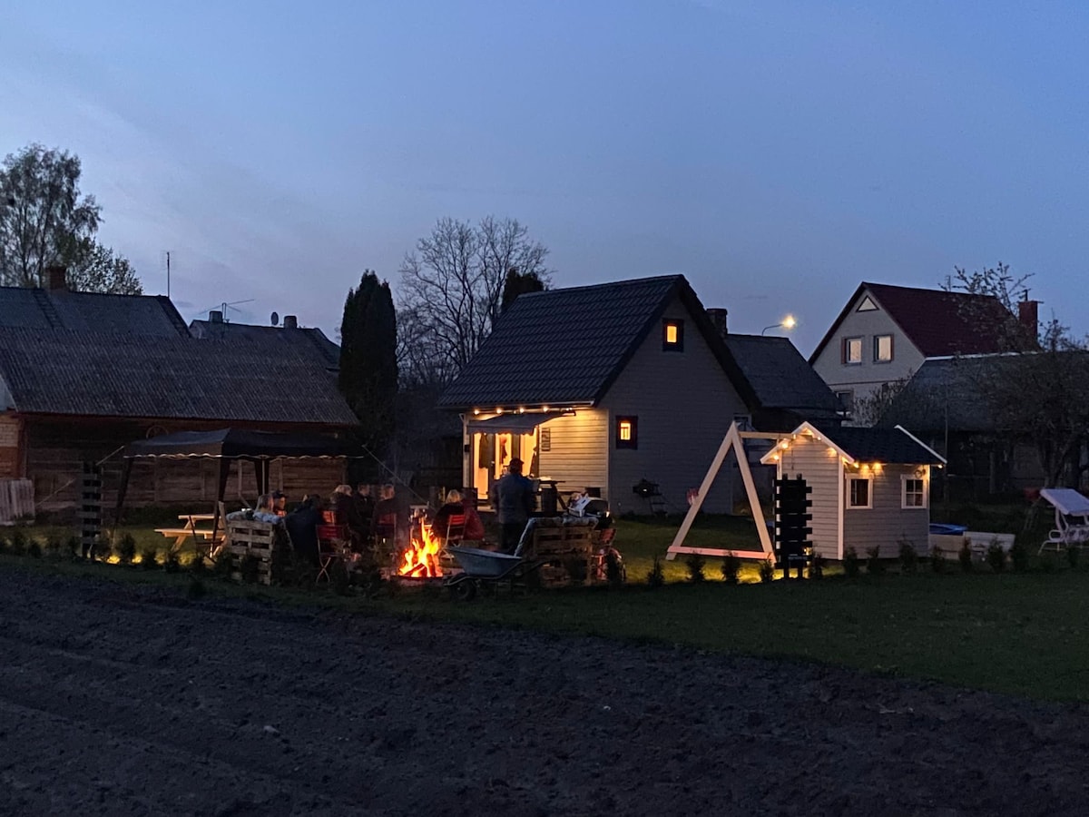 The exterior of the tiny house is illuminated at dusk, highlighting a cozy seating area with a fire pit. Nearby, a play structure is visible in the yard, surrounded by soft lighting and a peaceful atmosphere. Residential surroundings are gently integrated into the scene.
