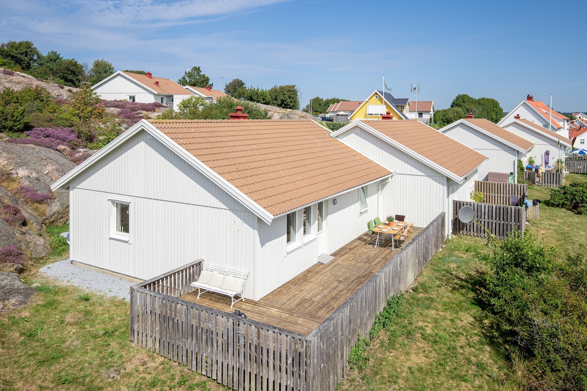A view of a single-story white home featuring a spacious wooden deck that extends across the front. The deck is furnished with a table and chairs, surrounded by a grassy area and partial fencing. Nearby houses and greenery create a pleasant neighborhood setting.