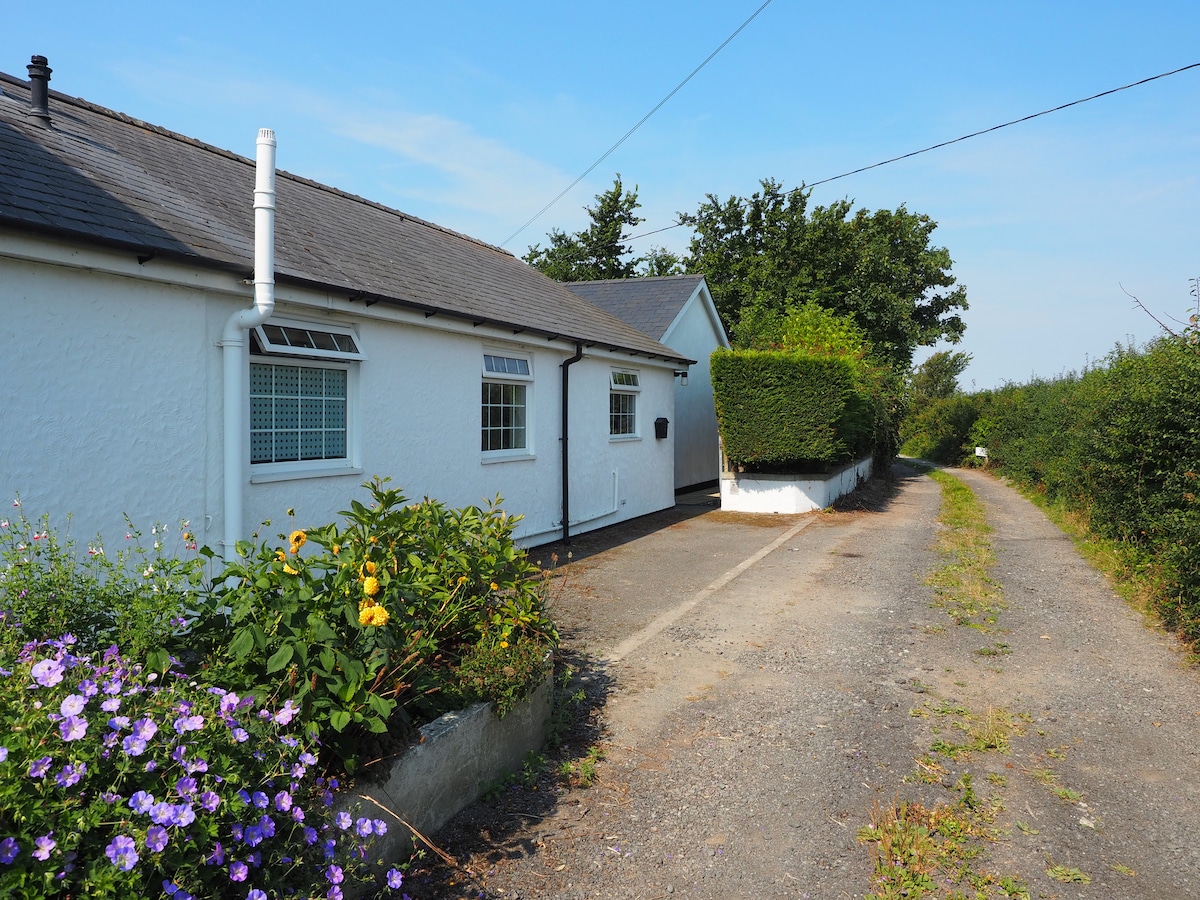 A white cottage is situated along a gravel pathway, bordered by well-maintained greenery and vibrant flowers. The building features multiple windows and a gently sloping roof, set against a clear blue sky and surrounded by open fields.