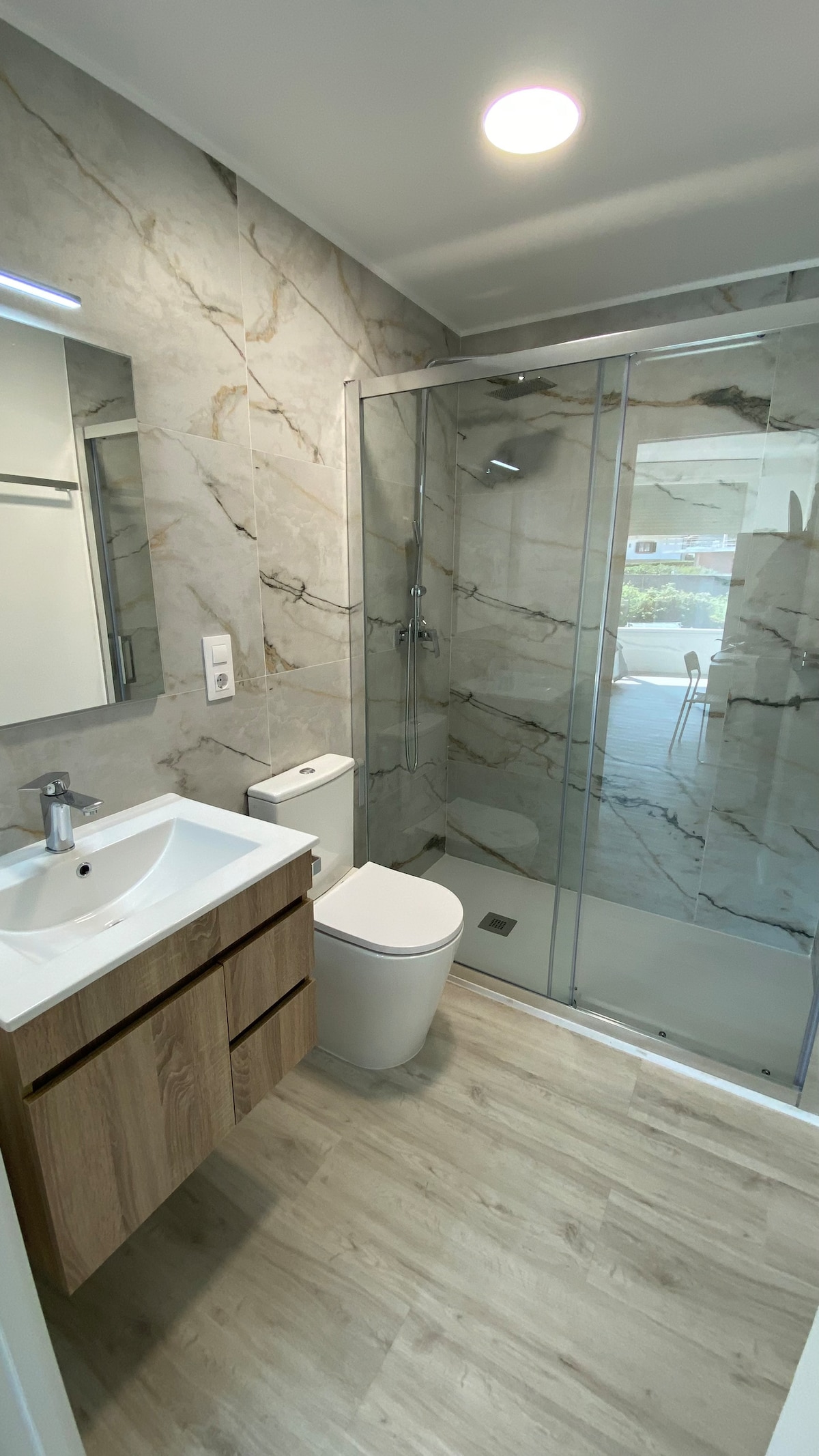A modern bathroom features a sleek glass shower with a rainfall showerhead. A wall-mounted mirror reflects the space, complemented by wooden cabinetry beneath a white countertop. Natural light filters in through a window, illuminating the light-colored flooring and neutral wall tiles.