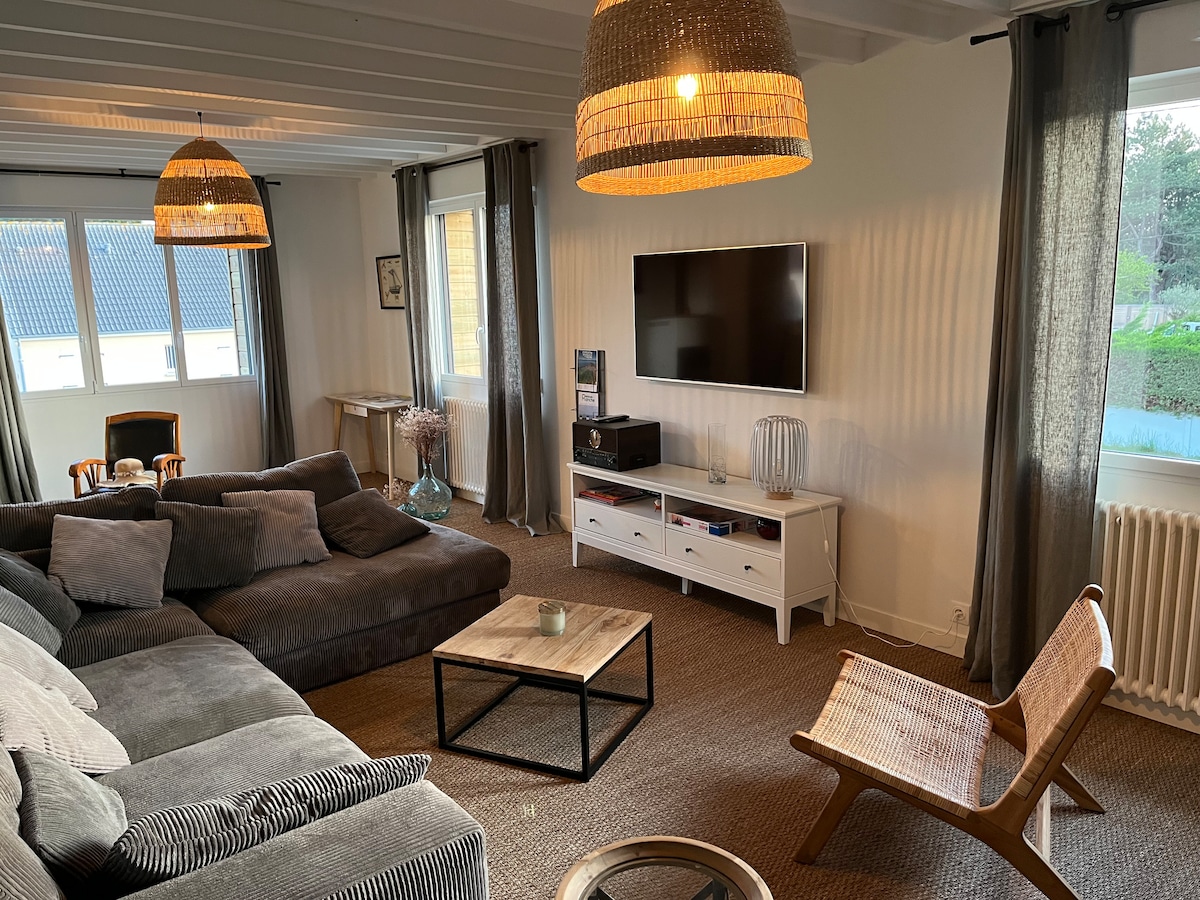The living area is characterized by a spacious sectional sofa in gray tones, accompanied by a light wood chair. A coffee table sits centrally, with a flat-screen TV mounted on the wall. Natural light enters through two large windows, enhancing the inviting ambiance.