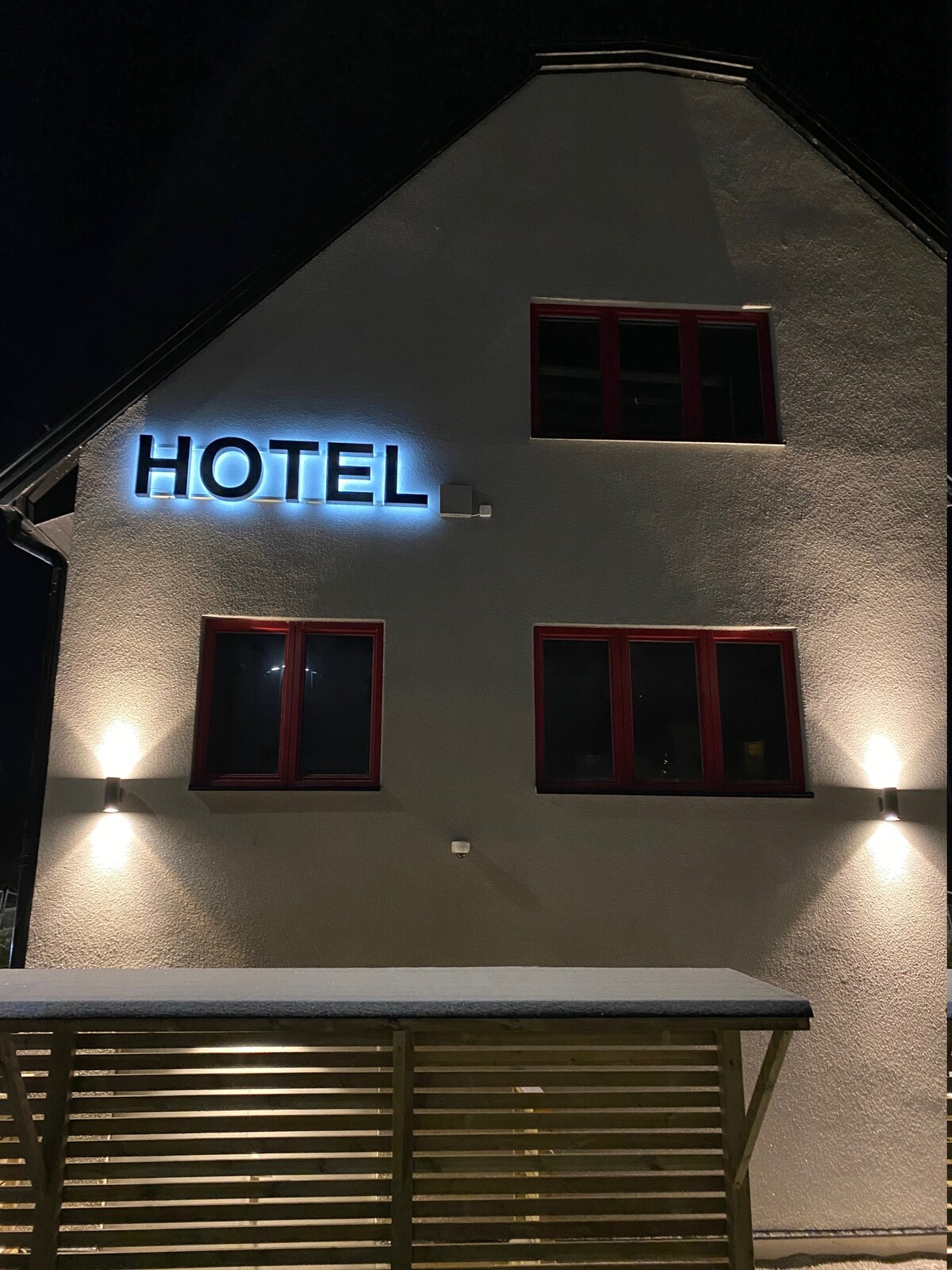 The exterior of the hotel is showcased at night, featuring a modern façade with illuminated signage displaying the word 'HOTEL.' Three windows are visible, with vibrant red frames adding a pop of color against the light-colored walls.