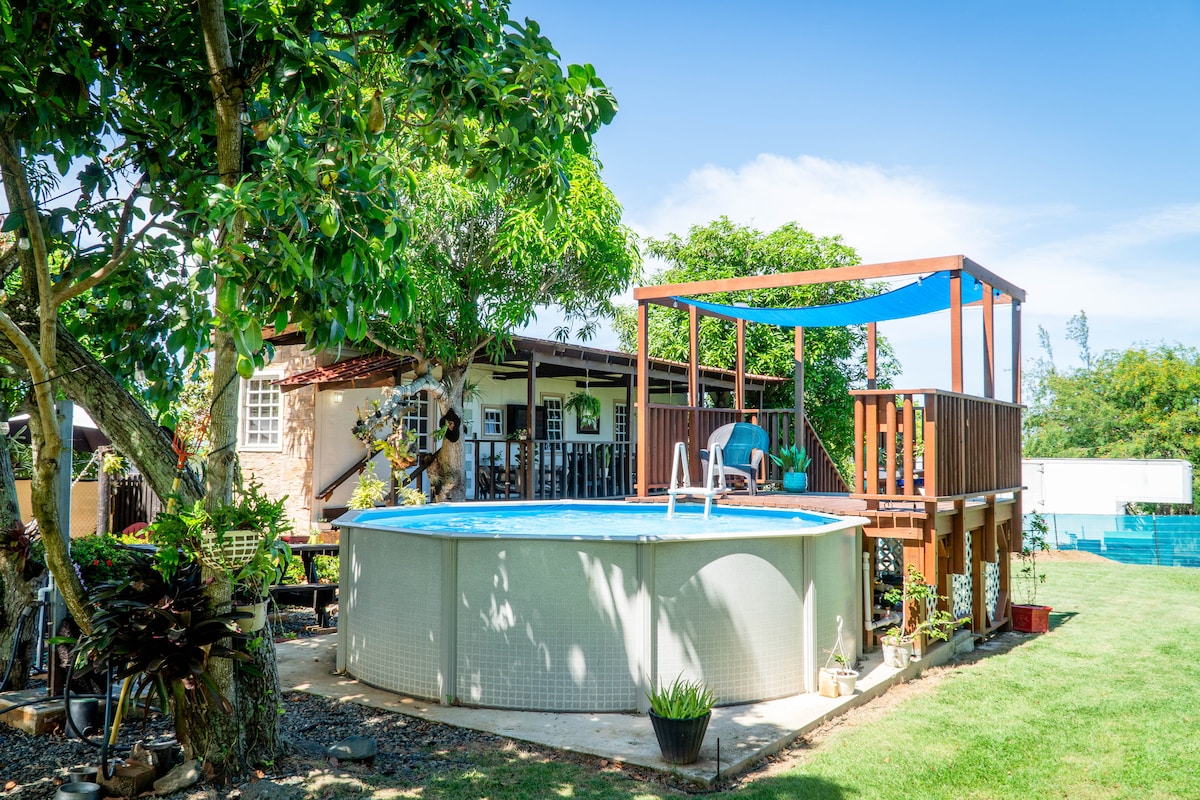A round pool is positioned next to a deck with shaded seating, surrounded by green grass and trees. The trailer home is visible in the background, featuring a patio area with comfortable furniture. Sunlight casts a warm glow on the outdoor space, creating a serene atmosphere.
