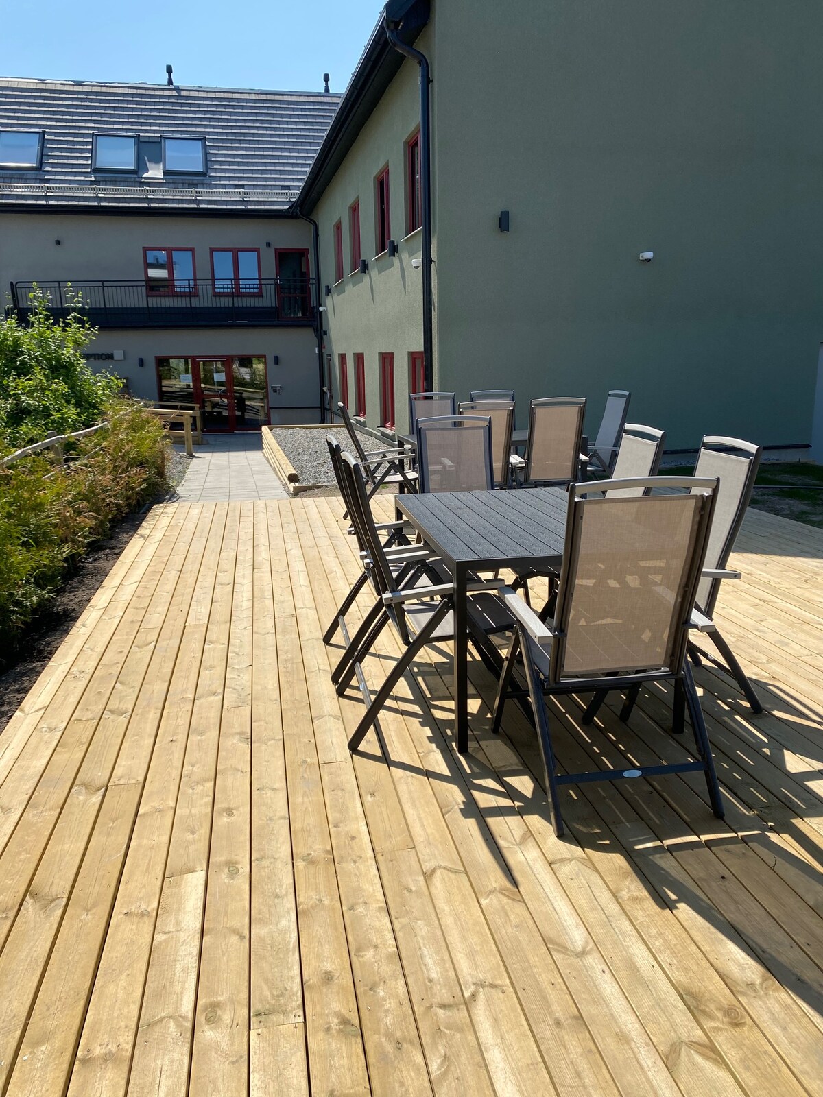 An outdoor seating area features a long table surrounded by several chairs on a wooden deck. The backdrop includes a multi-story building with large windows and a doorway leading inside. Lush greenery lines the path, creating a serene setting.
