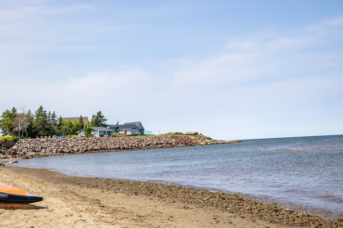 A sandy beach stretches along the tranquil shoreline leading to calm waters of Cocagne Bay. A rocky breakwater is visible in the distance with a collection of cottages surrounded by greenery. A clear blue sky complements the serene coastal scene.