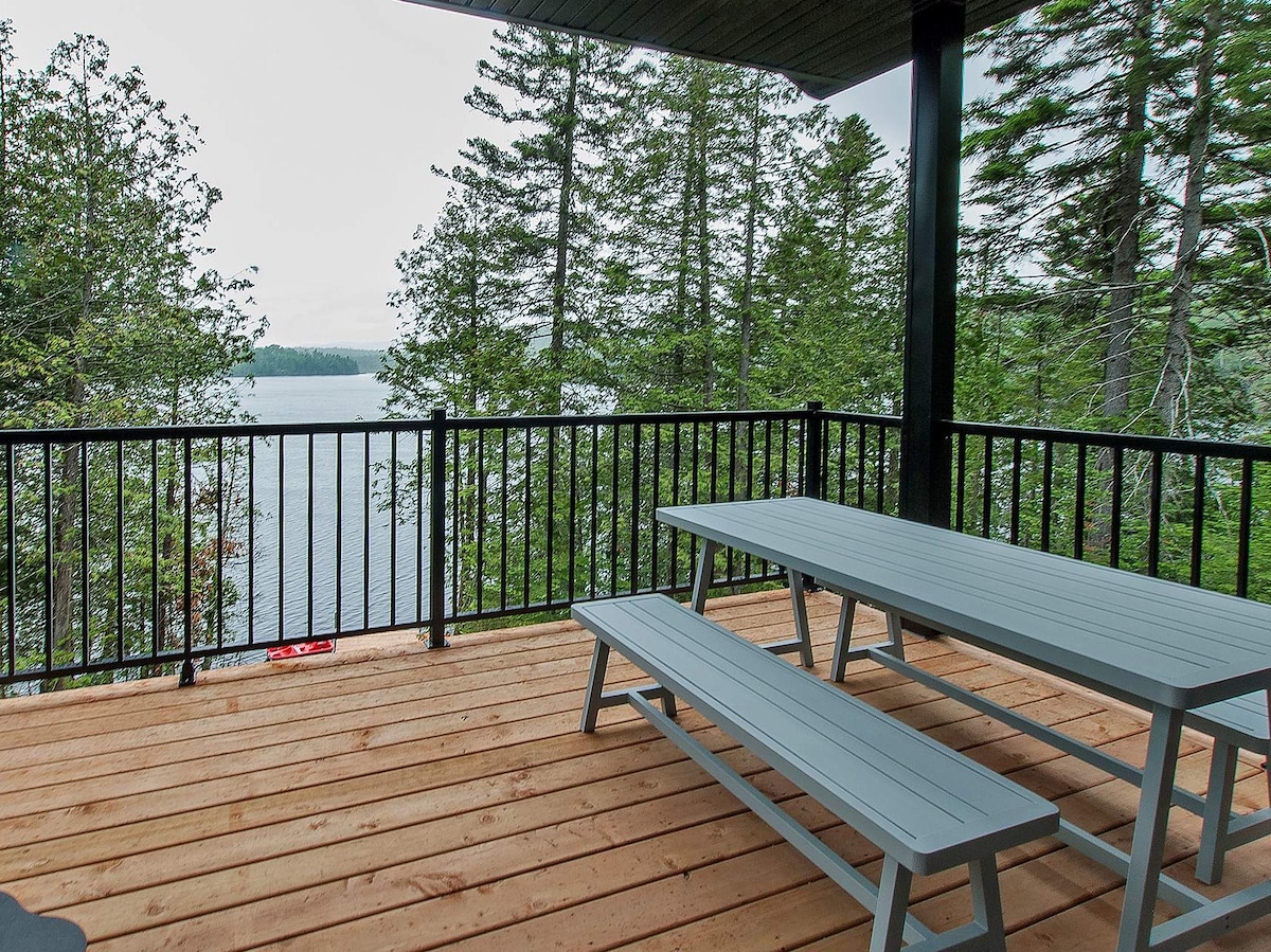 A wooden deck is viewed, featuring a picnic table with attached benches. The space is bordered by a black railing, allowing for an unobstructed view of the lake and surrounding trees, creating a peaceful outdoor setting.