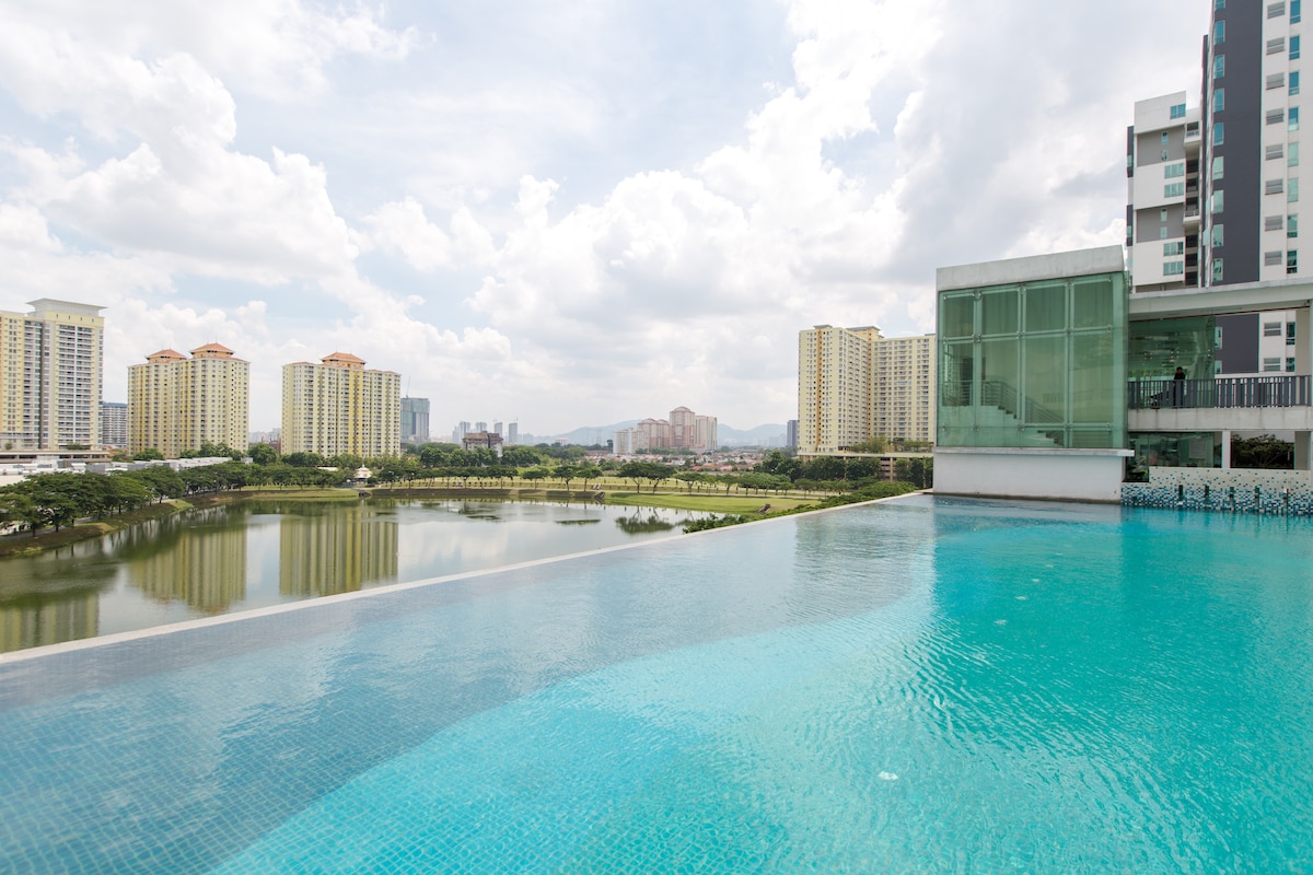 An expansive infinity pool is visible, extending towards a serene lake. Surrounding buildings reflect modern architecture, complemented by a clear sky and scattered clouds. The pool's water displays a bright turquoise hue, inviting relaxation and enjoyment.