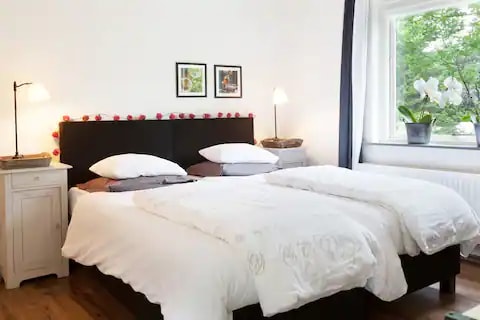A comfortable room features a spacious bed with white bedding and two pillows on each side. Soft lighting is provided by two bedside lamps, and a small side table is positioned next to the bed. A window offers a view of greenery outside, enhancing the natural light.