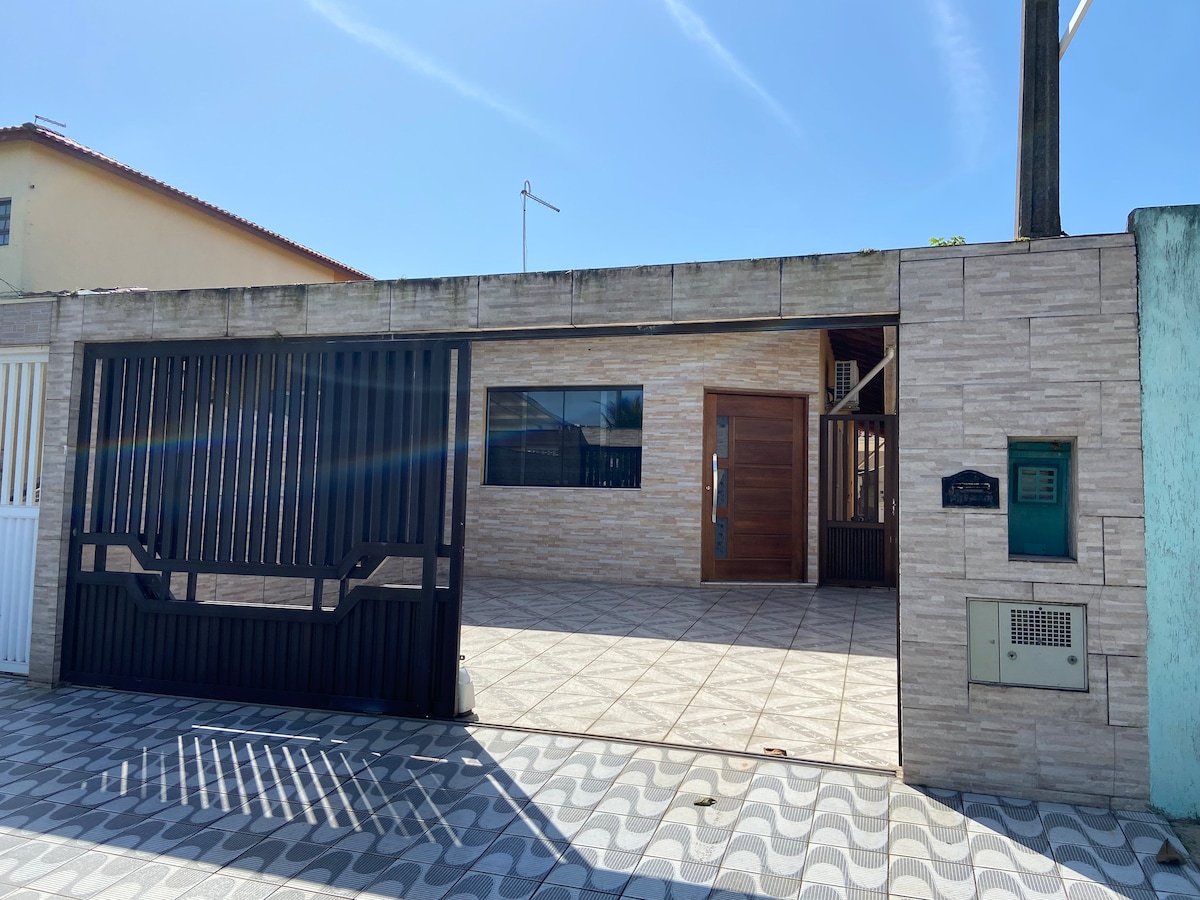 The exterior view features a modern one-story house with a stylish wooden door and large windows, allowing for natural light. A gated entrance with an automatic gate is visible. The surrounding area is paved, showcasing a clean and organized outdoor space.