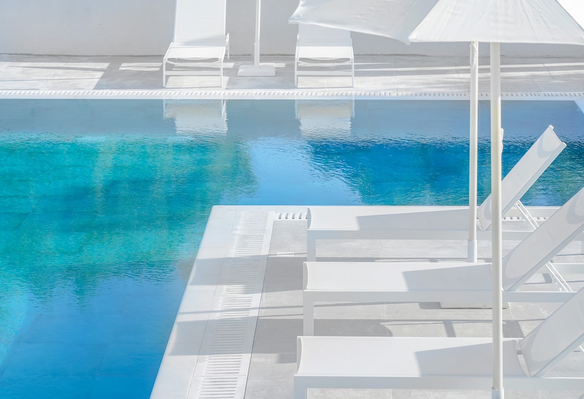 A tranquil outdoor pool is observed, featuring clear blue water reflecting sunlight. White lounge chairs are positioned around the pool area, accompanied by a large umbrella providing shade. The surrounding space is bright and well-maintained.