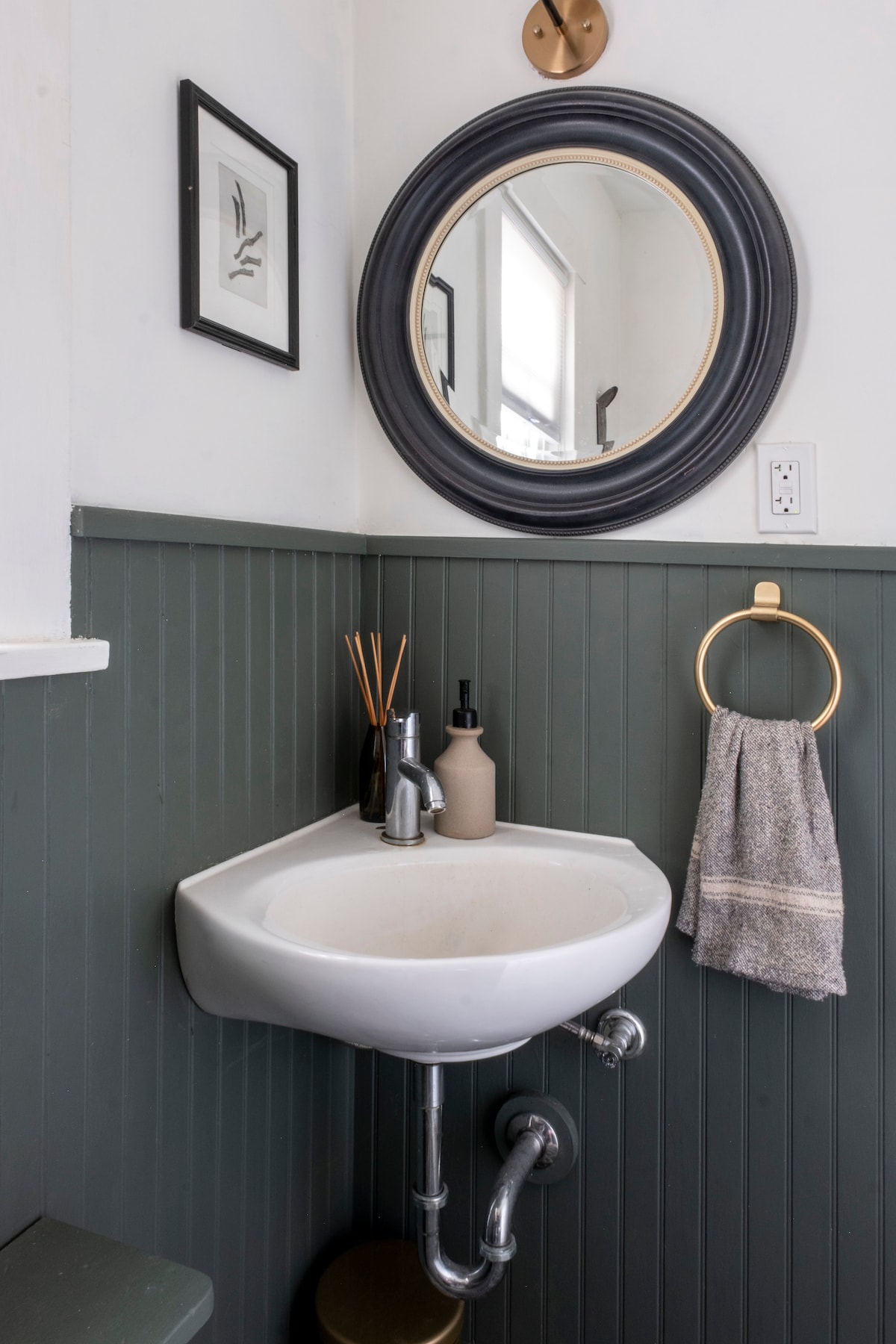 A compact bathroom corner features a modern sink with a sleek faucet, complemented by a round mirror framed in black. The lower half of the wall is painted dark green, with a hand towel hanging on a gold ring and decorative items placed nearby.