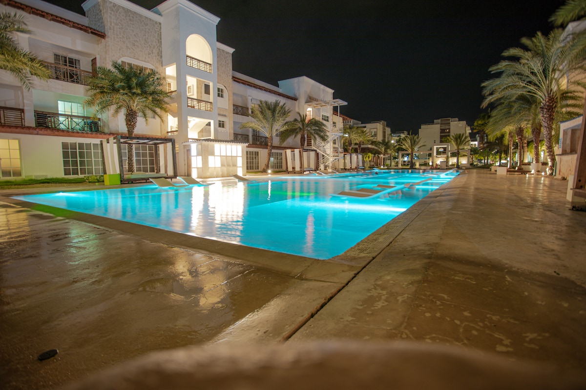 A tranquil outdoor pool area is illuminated by soft blue lighting at night. Surrounding palm trees add a tropical touch, while neighboring buildings provide a spacious community setting. The clean, smooth surface of the pool reflects the light, creating a serene atmosphere perfect for relaxation.