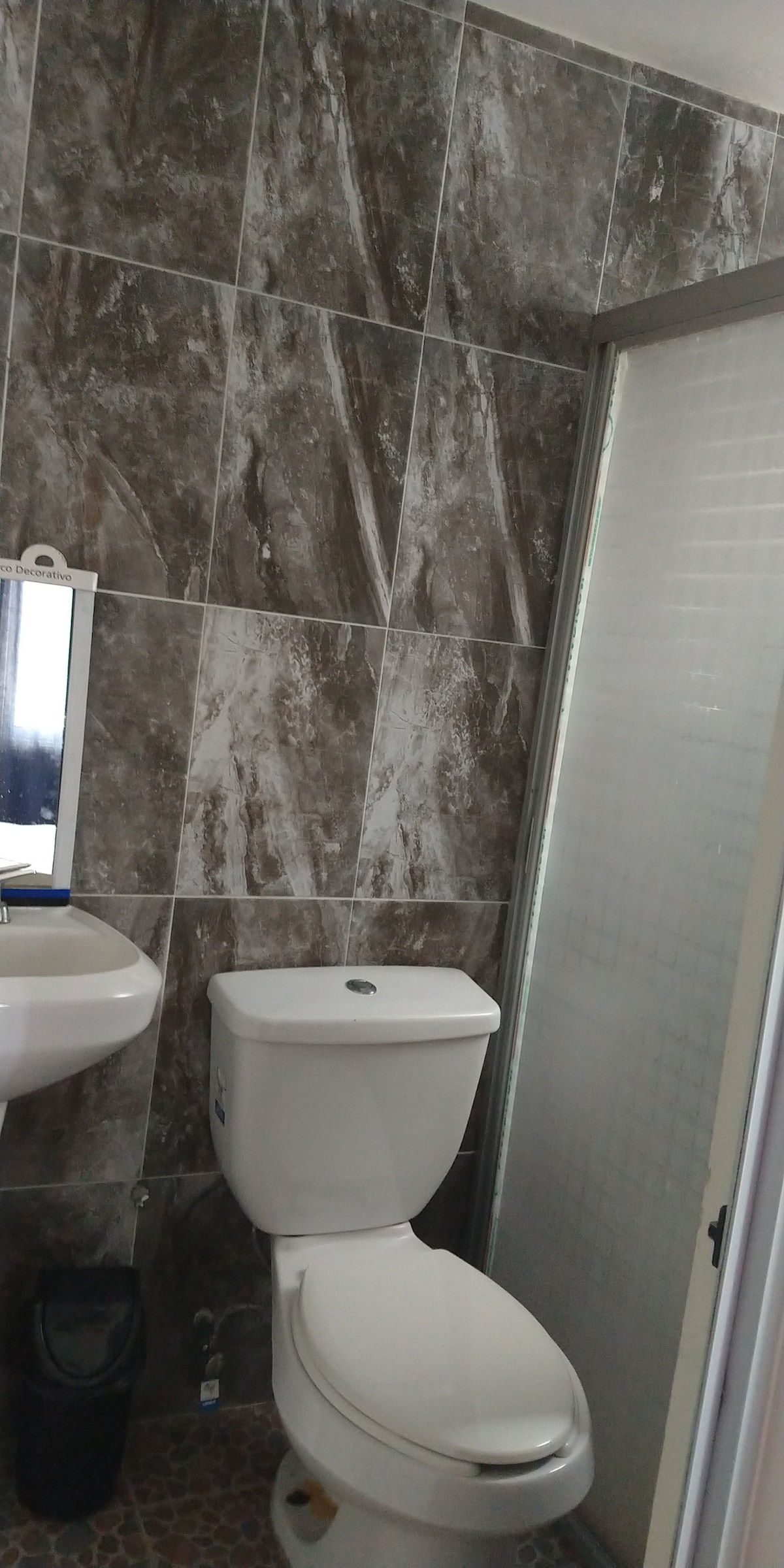 A compact bathroom features modern stone-textured walls. A white toilet and a white sink with a mirror above are visible. A glass shower enclosure complements the space, with light filtering through the frosted glass.