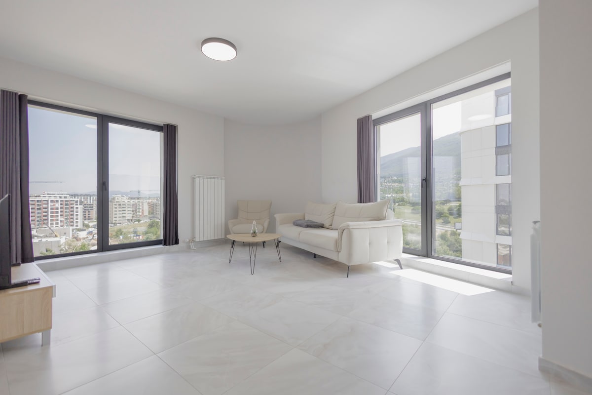 A bright and spacious living area features large windows offering panoramic views. A comfortable sofa is complemented by a small coffee table, creating an ideal space for relaxation. Modern light fixtures enhance the open layout, while natural light fills the room.