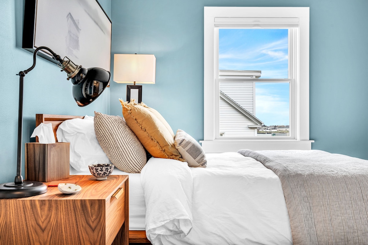 A bright bedroom features a queen bed with soft white linens and decorative pillows in warm tones. A wooden nightstand holds a small bowl and a lamp. Natural light enters through a window, illuminating the calming blue walls.